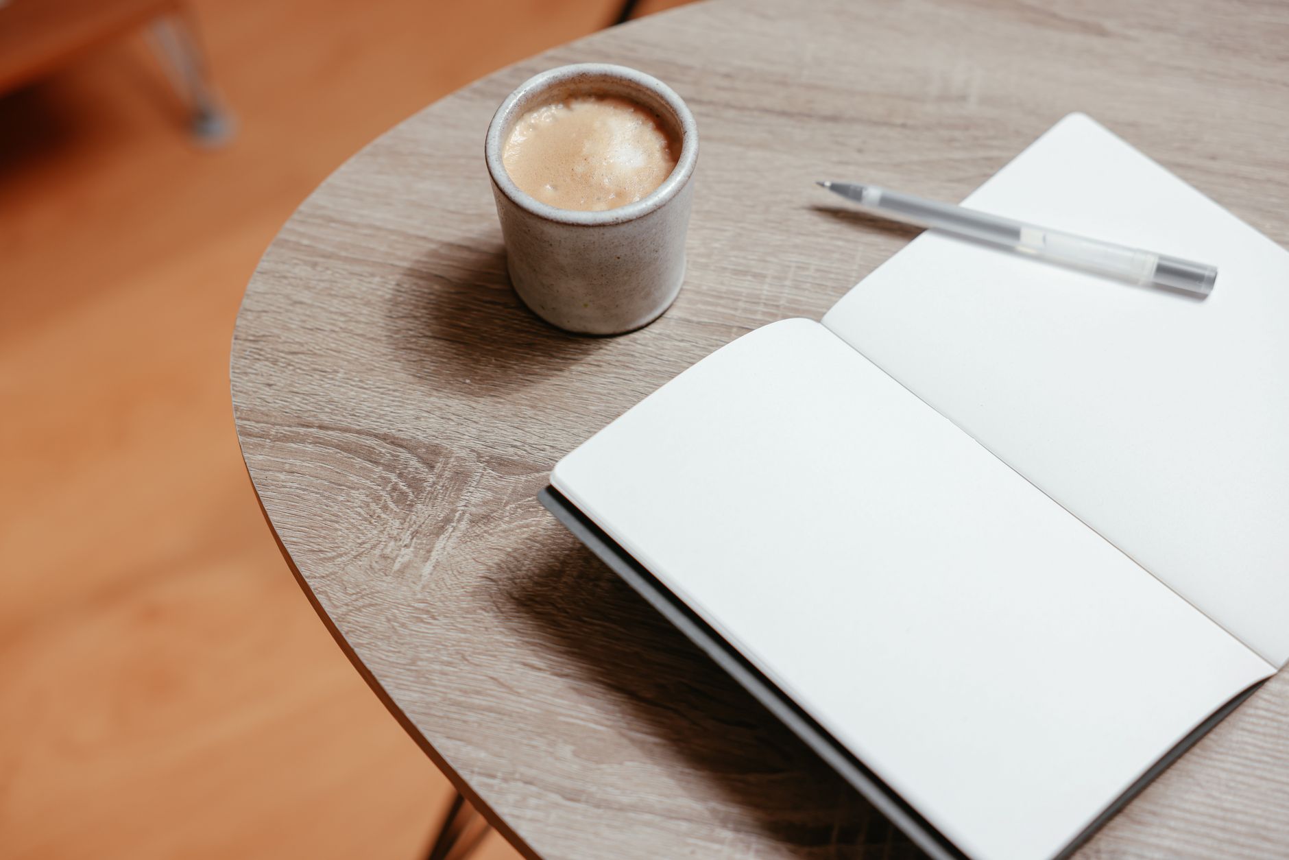 Carnet ouvert aux pages vierges avec un stylo, à côté d’une tasse en céramique, sur une table ronde en bois clair.